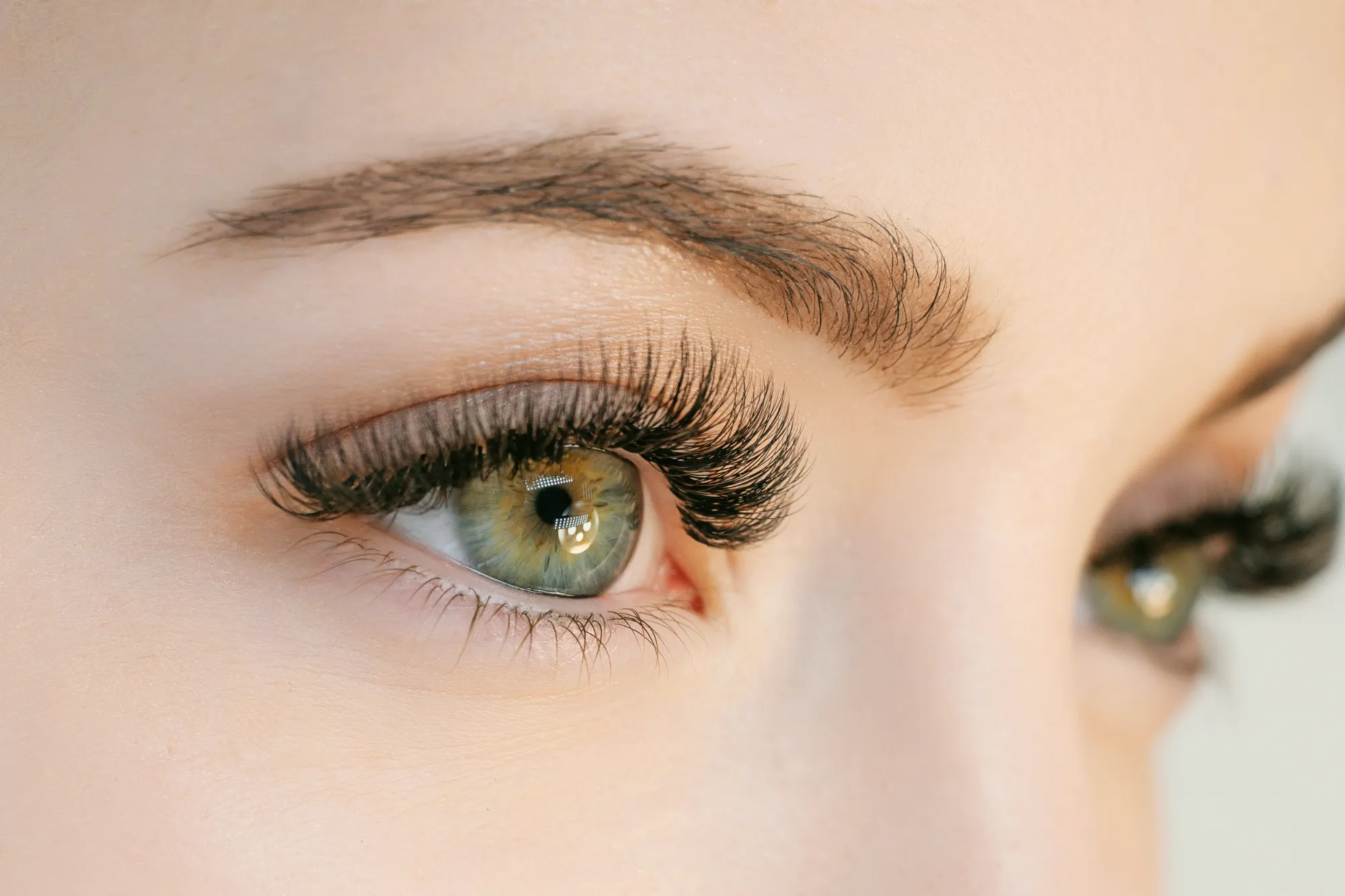 An extreme close-up of a bright green eye featuring dramatic, long, dark eyelash extensions and a well-groomed eyebrow.