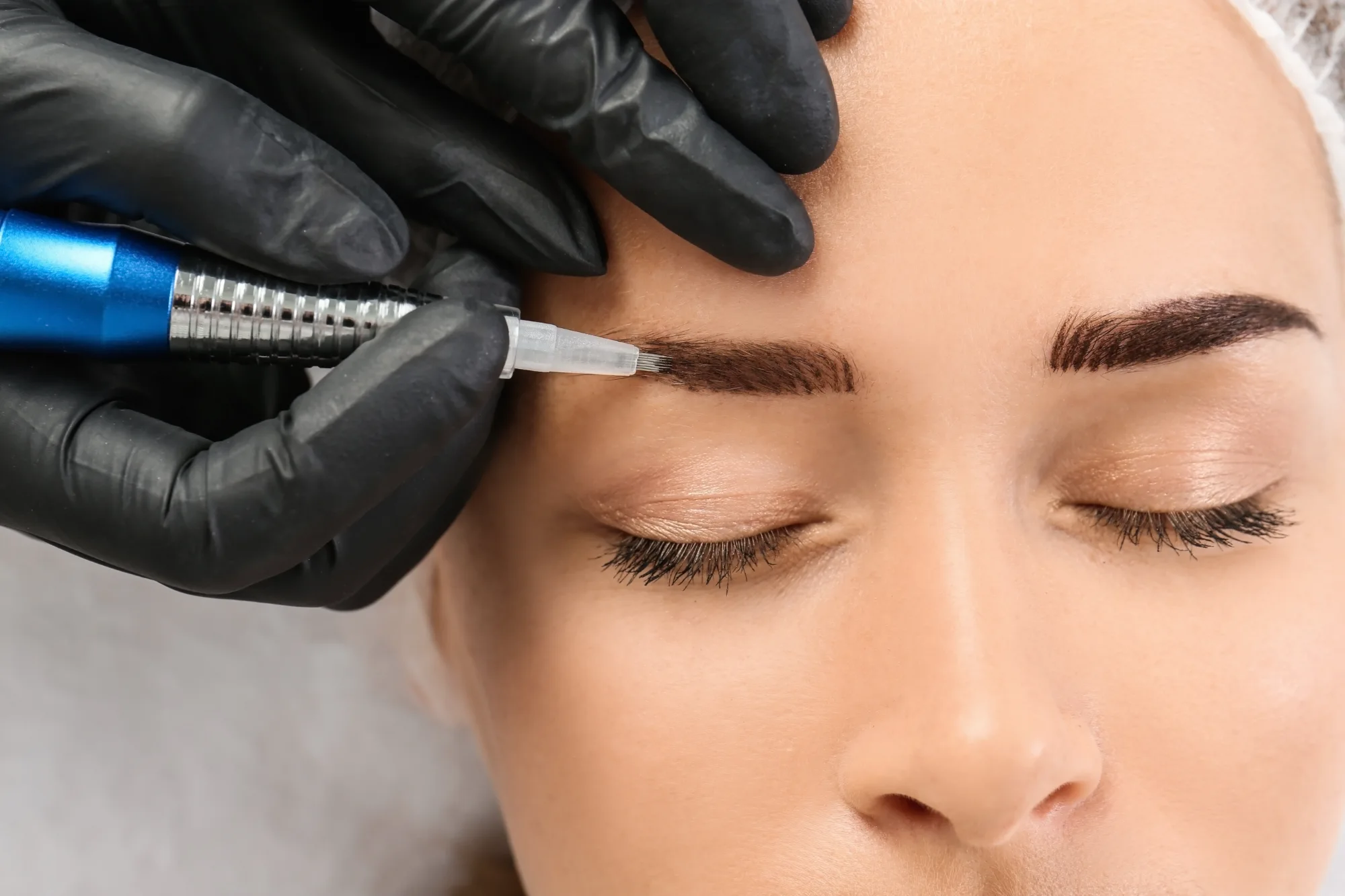 A close-up of a technician's hand wearing a black glove performing microblading on a client's eyebrow using a blue cosmetic tattoo pen.
