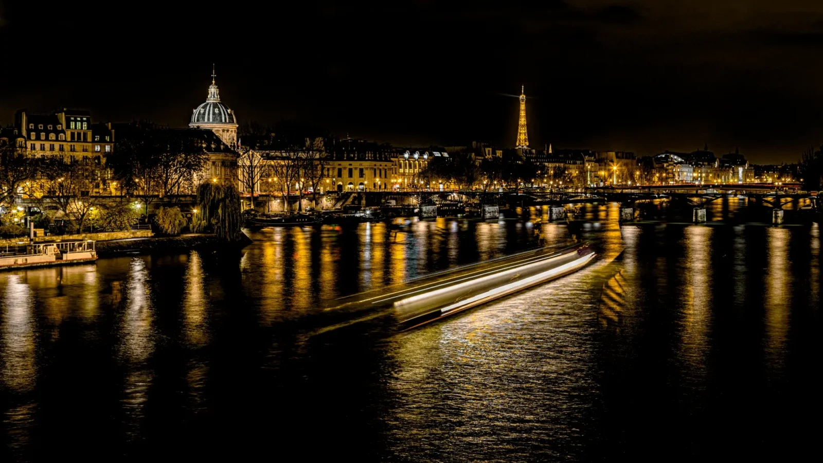 An illuminated Paris cityscape at night across the Seine River. The Eiffel Tower and the Institut de France dome are visible, while a boat blurs into a white streak on the water.