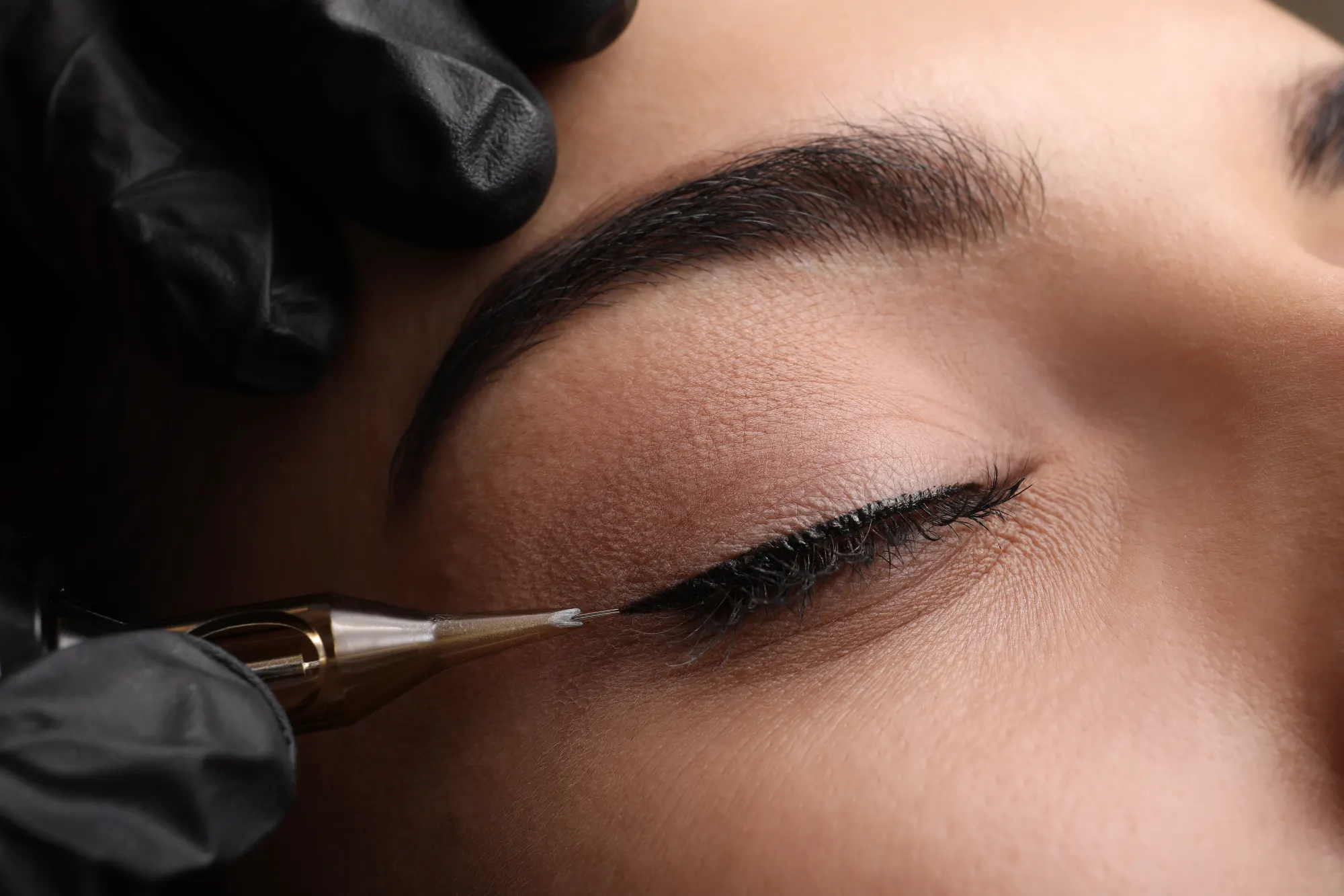 A close-up shows a technician wearing a black glove applying permanent eyeliner to a client's closed eye using a golden cosmetic tattoo pen.