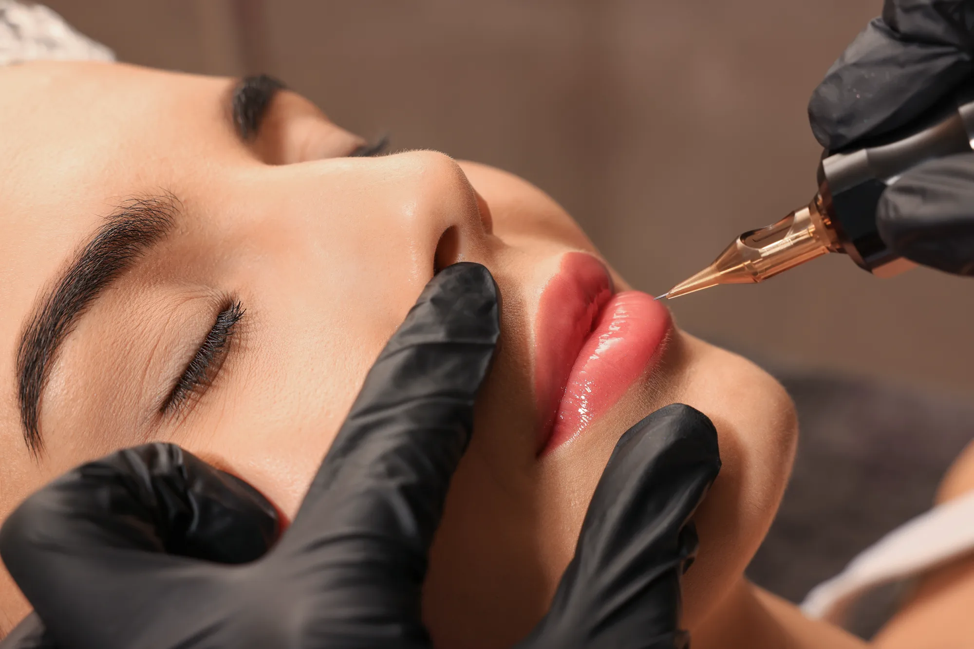 A close-up shows a client receiving permanent lip color applied by a technician wearing black gloves, who is using a small tattooing device.