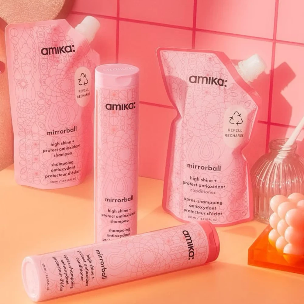 Four pink Amika "mirrorball" hair care products, including shampoo and conditioner bottles and refill pouches, sit on a surface against a pink tiled wall.