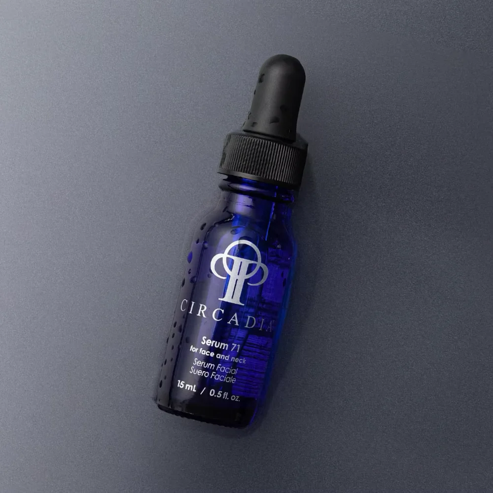 A dark blue glass bottle of Circadia Serum 71 for the face and neck, featuring a black dropper top, rests diagonally on a dark gray surface.