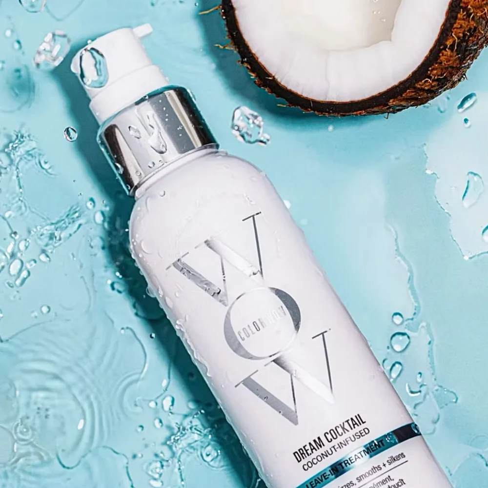 A white Color Wow hair treatment bottle (Dream Cocktail Coconut-Infused) sits on a wet, light blue surface with a halved coconut beside it.