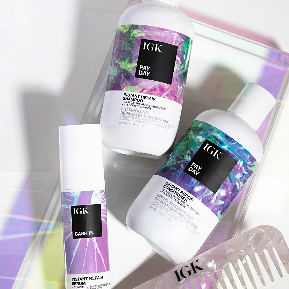 Three IGK hair care products, including Pay Day shampoo, conditioner, and Cash In serum, are displayed on a shimmering tray. The labels feature abstract purple and green designs.