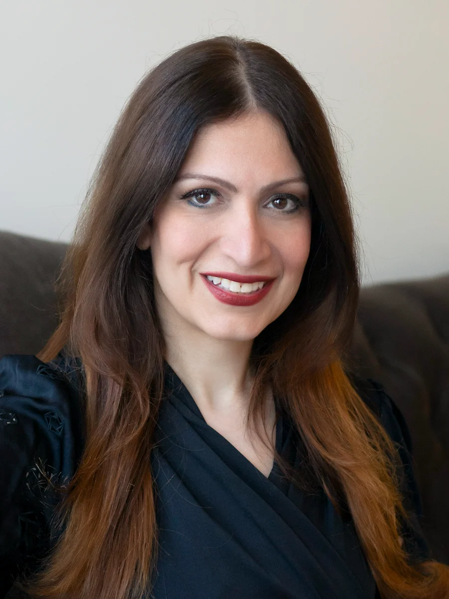 Brigitte Rist, the salon owner and stylist, smiles warmly in a professional portrait. She has long, dark hair and wears a black top.