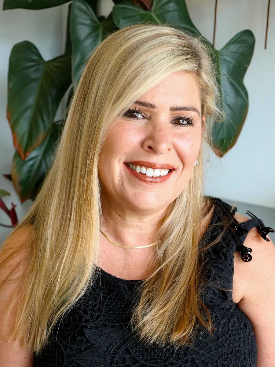Stylist Renee Wojtylo with blonde hair smiles warmly. She is wearing a black lace top and a thin gold necklace, standing in front of large green plant leaves.