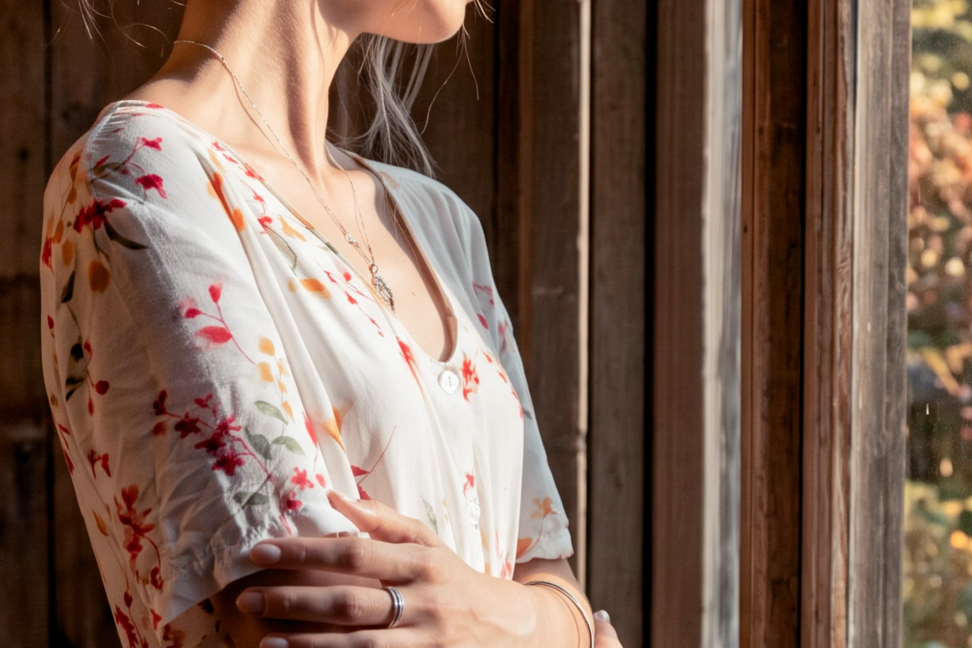 Close-up of a woman in a white floral print dress and necklace standing by a rustic window in warm sunlight.
