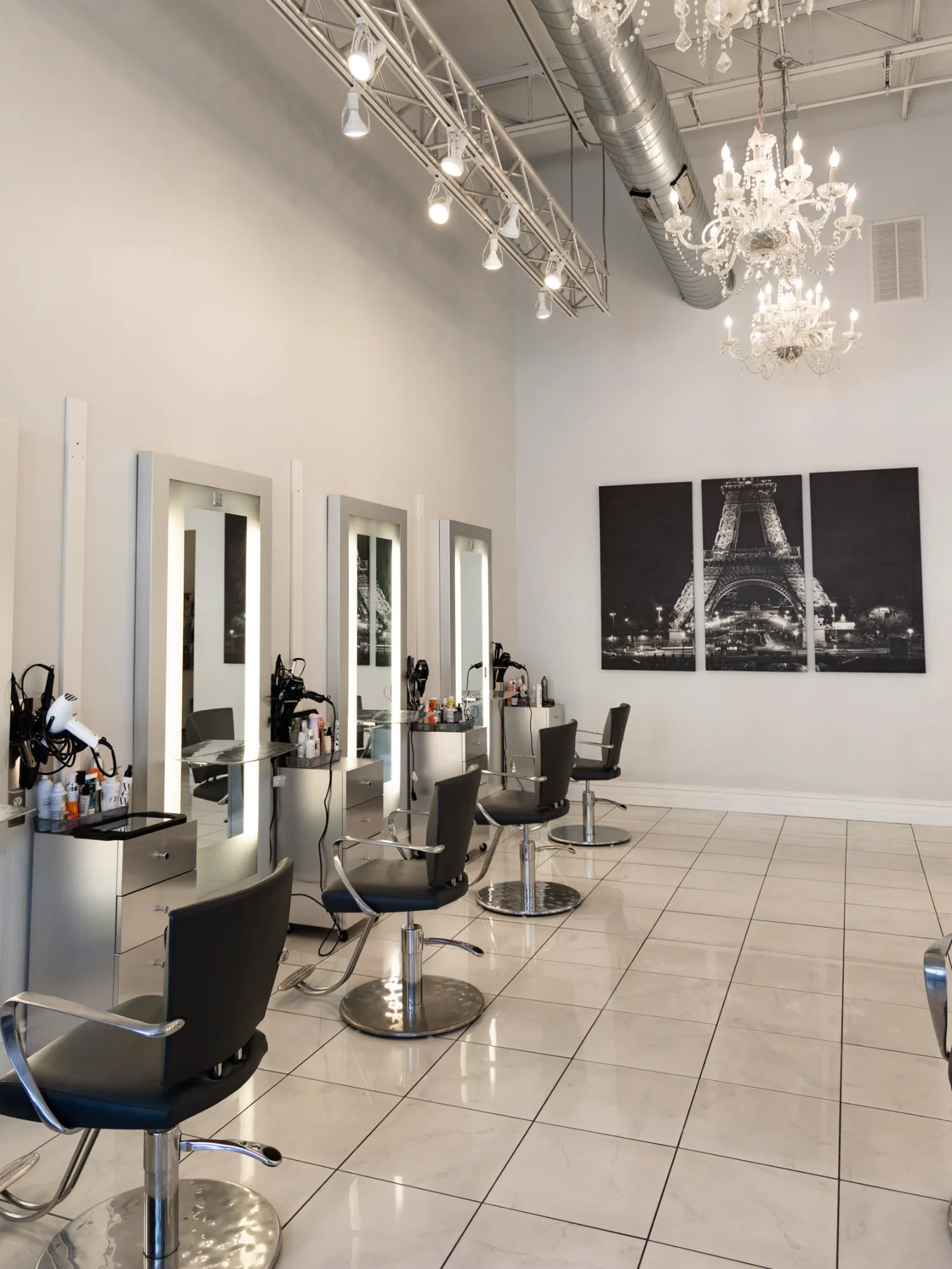 An upscale beauty salon interior featuring three lighted stylist stations, black chairs, white tile floors, and dramatic crystal chandeliers hanging near a large Eiffel Tower triptych.