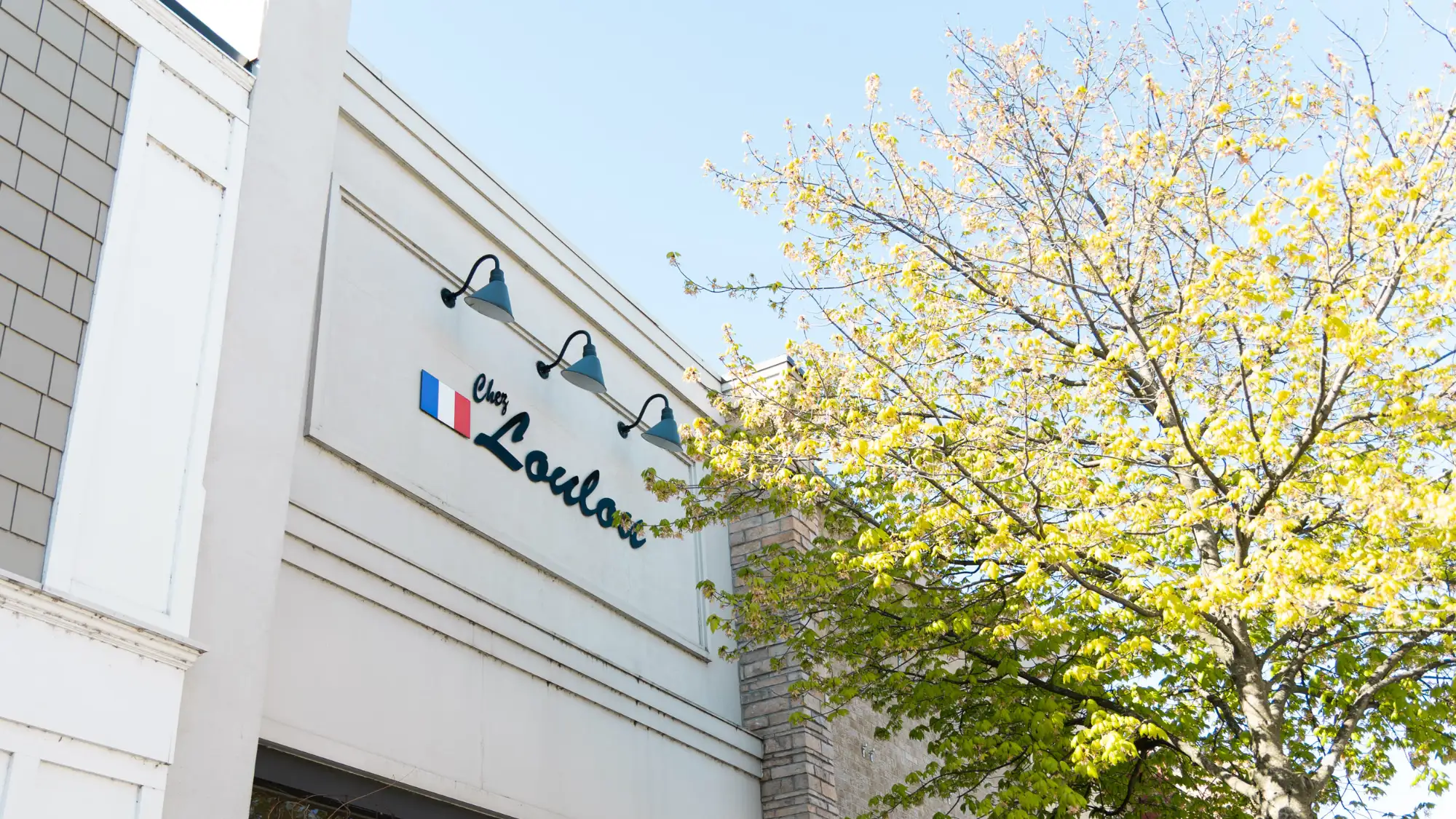 The exterior sign for Chez LouLou, featuring a French flag symbol, is mounted on a white wall under three lights. A large tree with bright spring foliage is to the right.