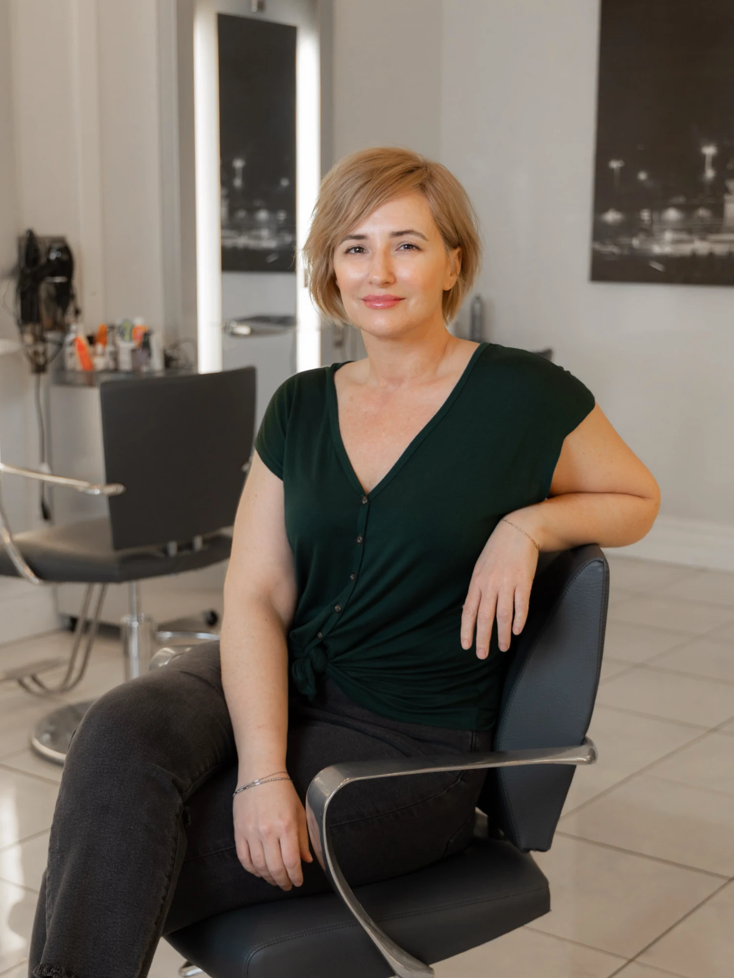 Esthetician Misty Balido sits casually in a black salon chair, wearing a dark green top and black jeans, smiling confidently at the viewer in the salon environment.