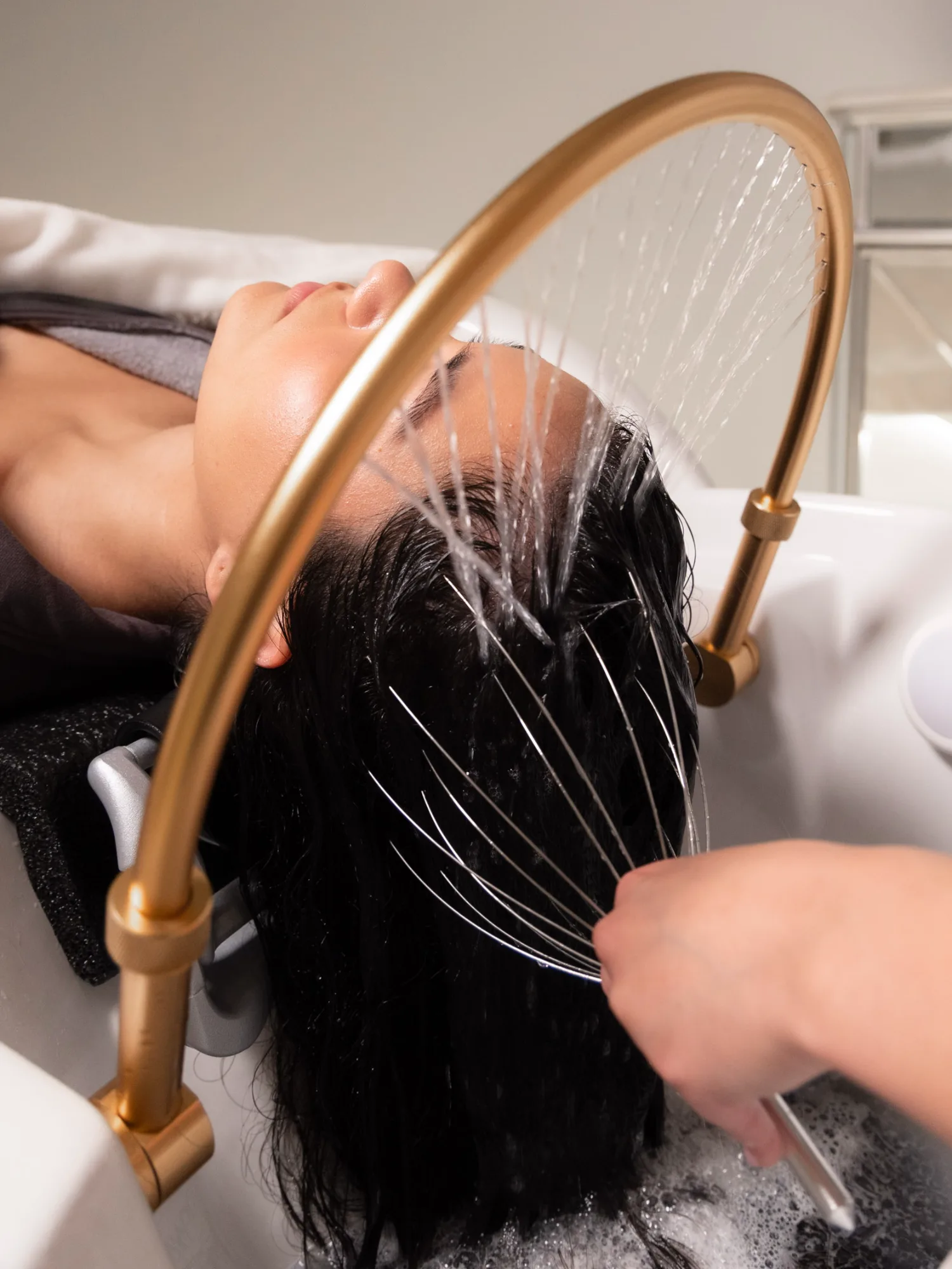 A client with dark hair lies in a basin, receiving a Japanese head spa treatment. Water flows from a gold fixture as a hand uses a wire scalp massager.