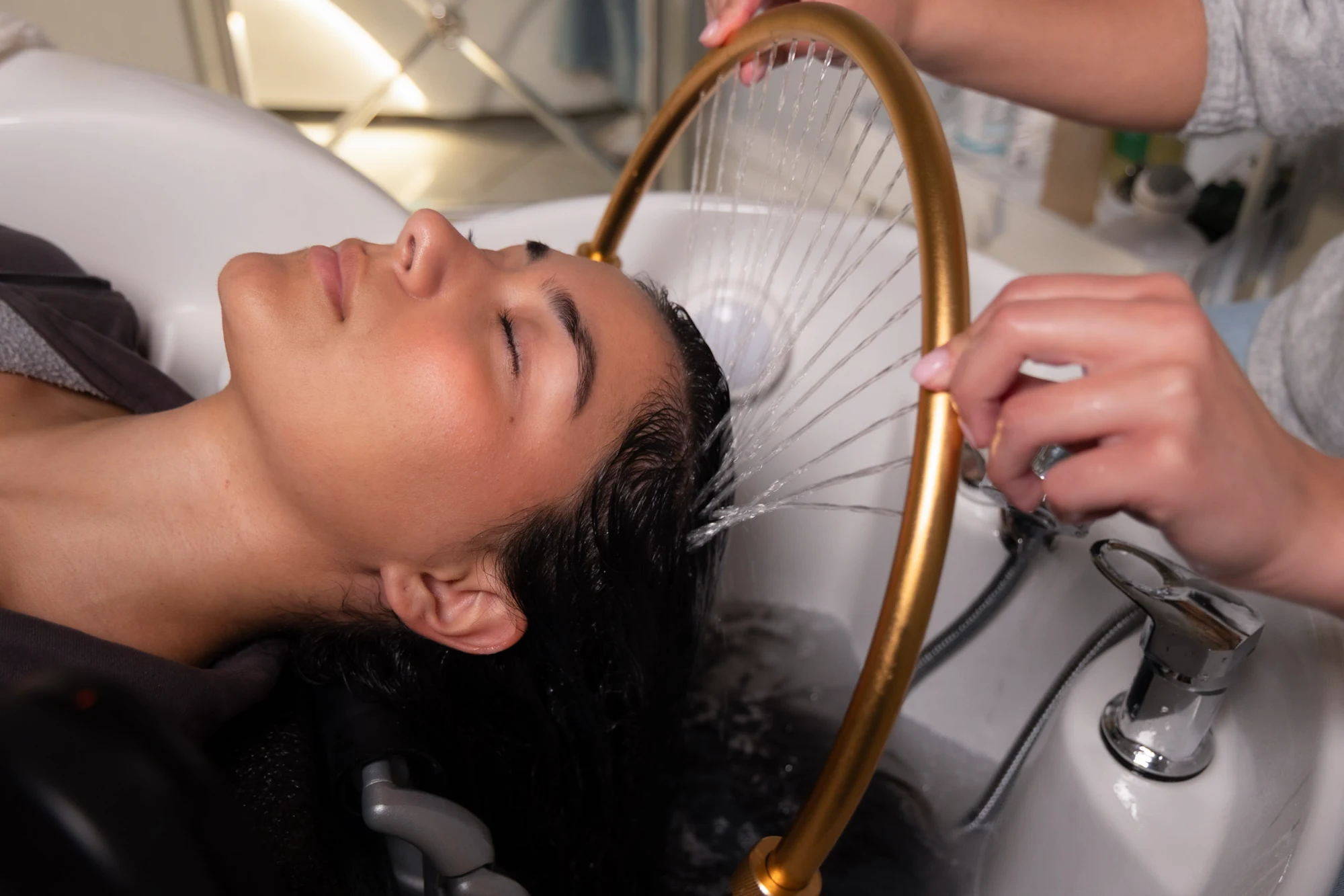 A client with dark hair lies back in a white salon basin, eyes closed, receiving a Japanese head spa treatment with water streaming from a gold circular fixture.