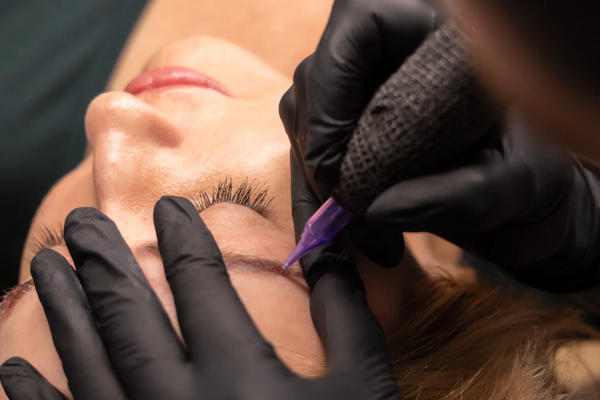 Lindsay, a technician, uses a nano-technique tattoo pen to carefully apply permanent makeup to a client's eyebrow while wearing black gloves.