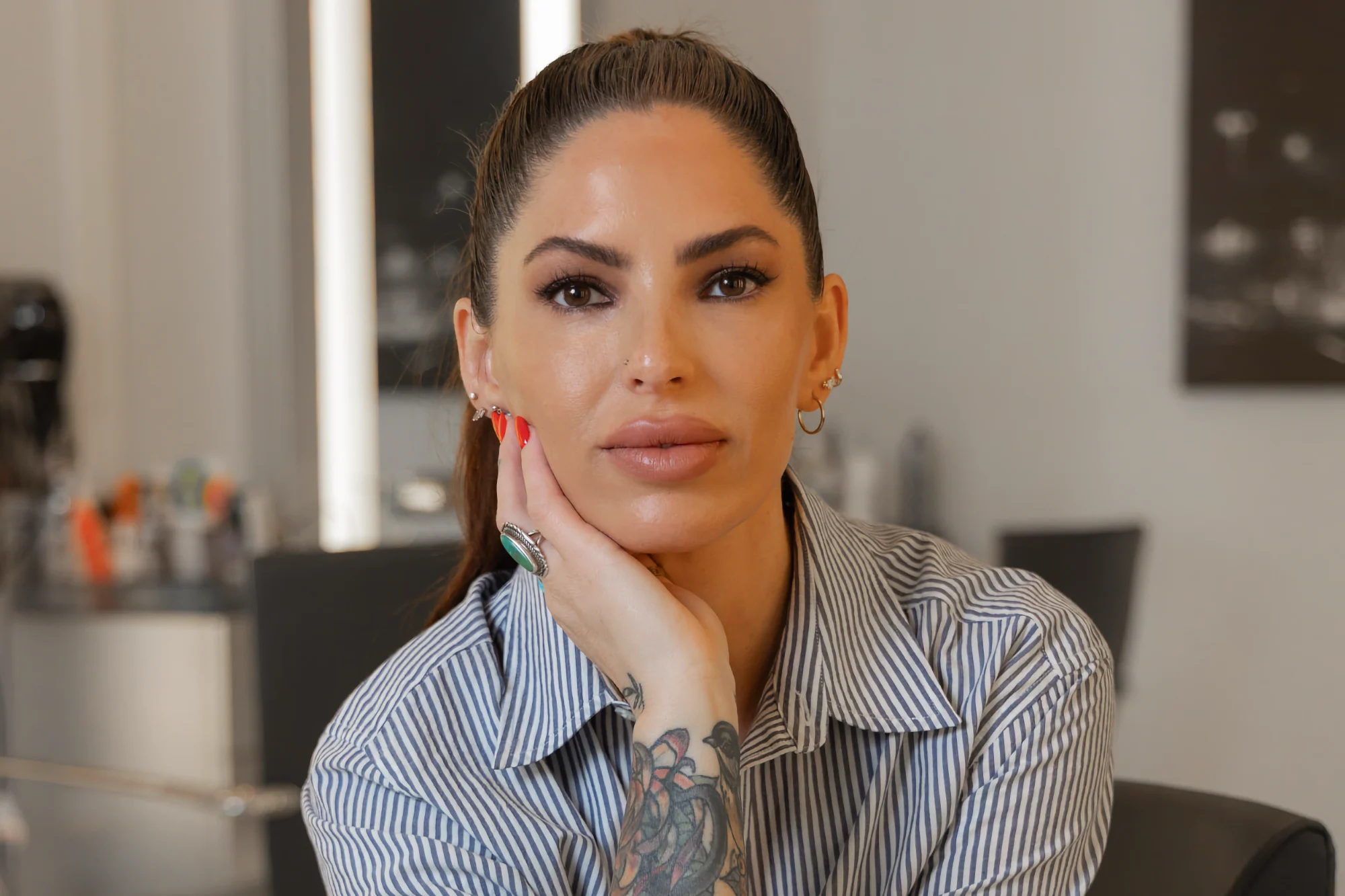 Lindsay Trudell, a permanent makeup tattoo artist, looks directly forward. She wears a striped shirt and rests her chin on her hand, showing arm tattoos.