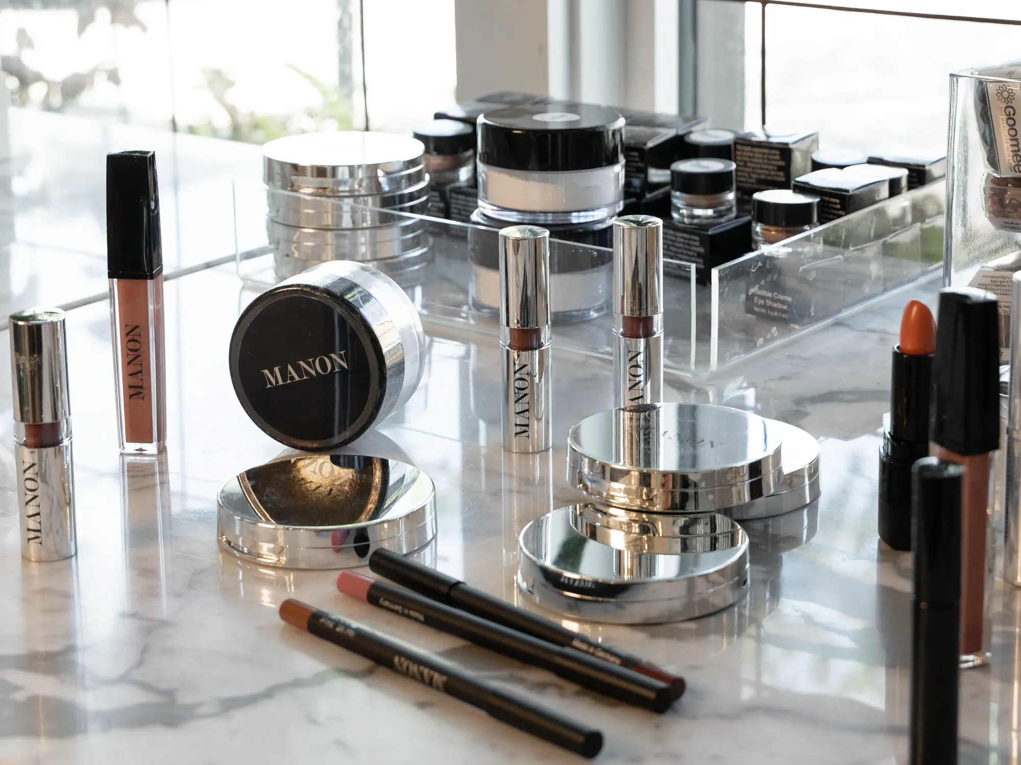 A selection of Manon beauty products, including silver compacts, lipsticks, and lip liners, displayed on a polished white marble counter.