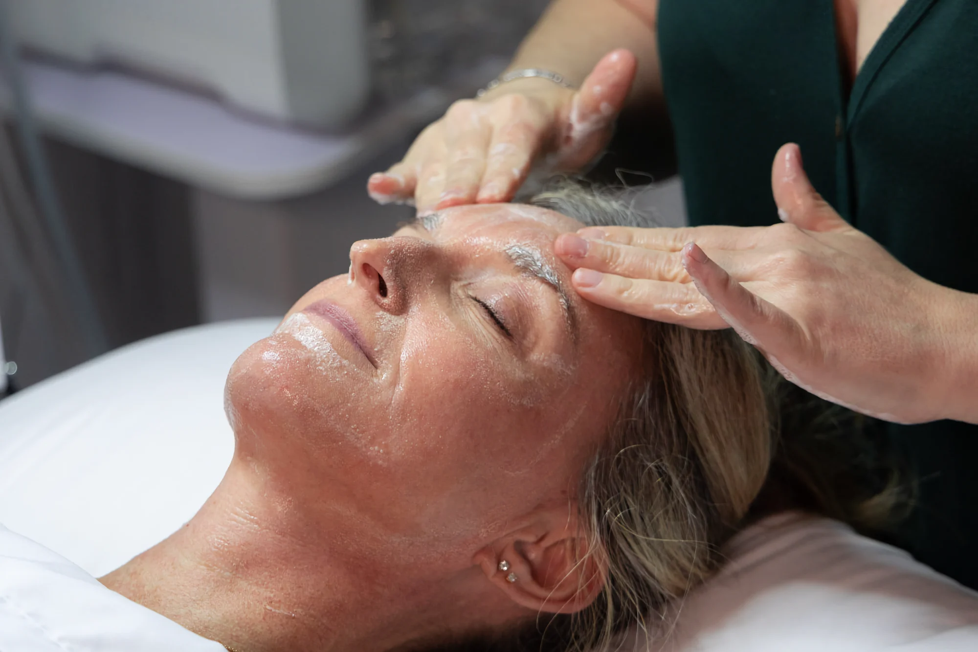 A female client lies relaxed with eyes closed while aesthetician Misty Balido applies a facial treatment, massaging the creamy product into her forehead.