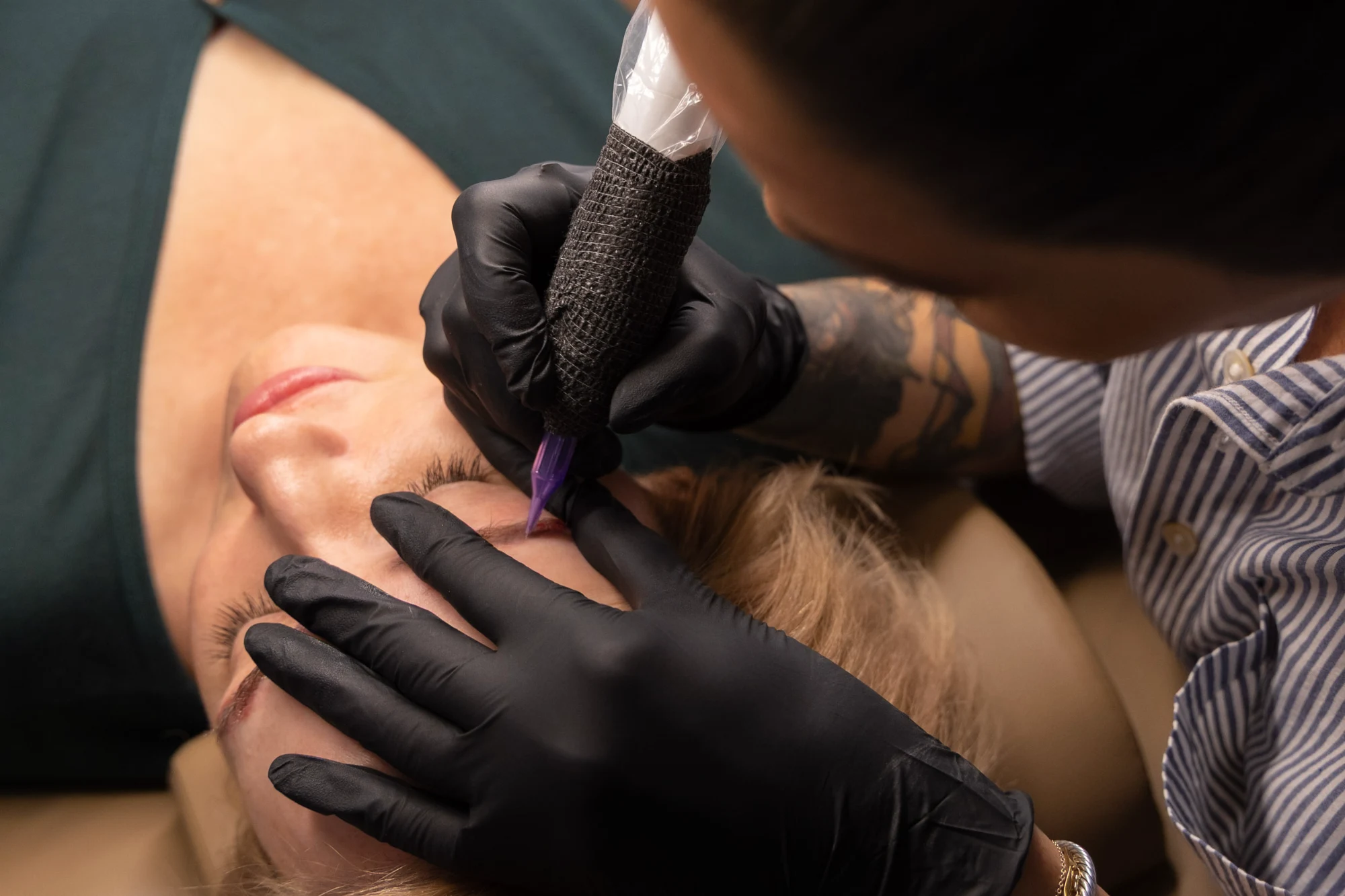 Lindsay Trudell wearing black gloves and a striped shirt applies a nano-brow treatment to a blonde client's eyebrow using a tattoo pen.