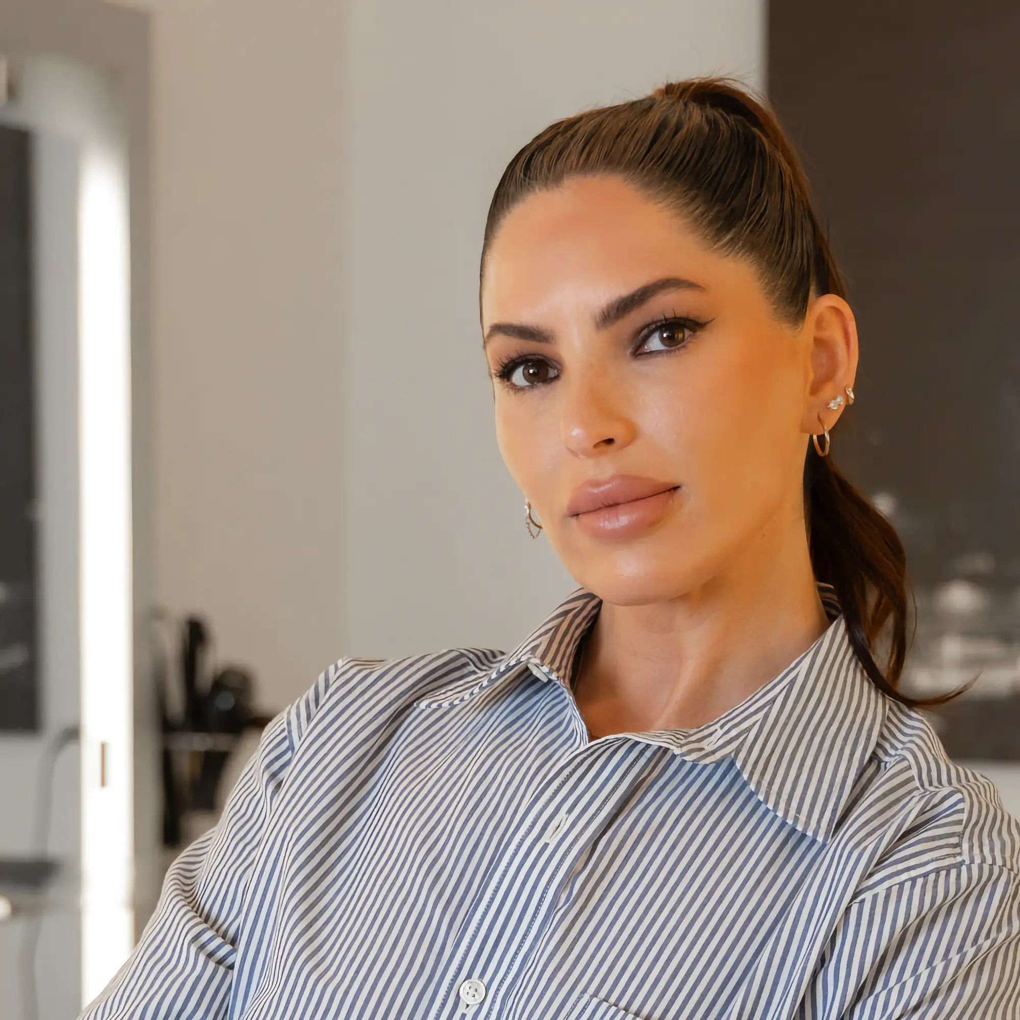 Permanent makeup artist Lindsay Trudell looks confidently at the camera. She wears a striped blue and white shirt and her dark hair is styled in a ponytail.
