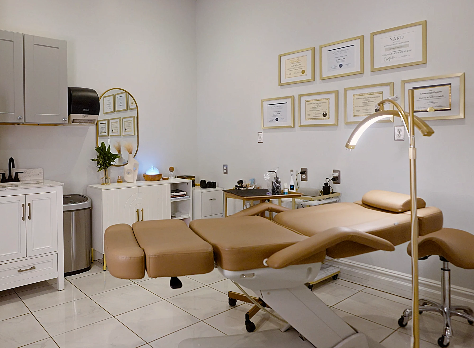 A bright, clean permanent makeup procedure room with a beige treatment chair, an arched gold lamp, and framed credentials on the white walls.