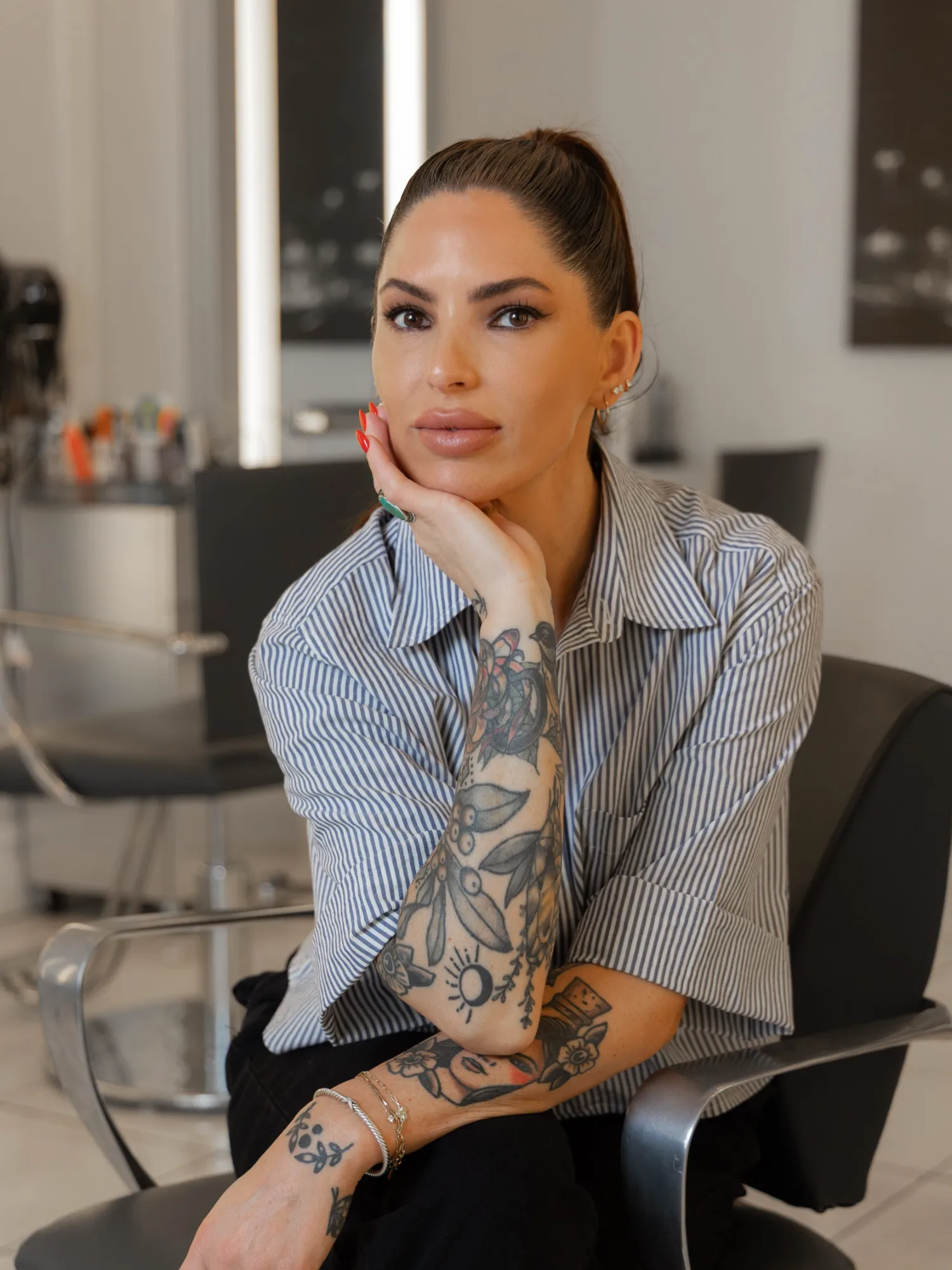 Permanent makeup artist Lindsay Trudell sits in a styling chair, looking forward. She wears a striped shirt, has full tattooed sleeves, and holds her chin thoughtfully.