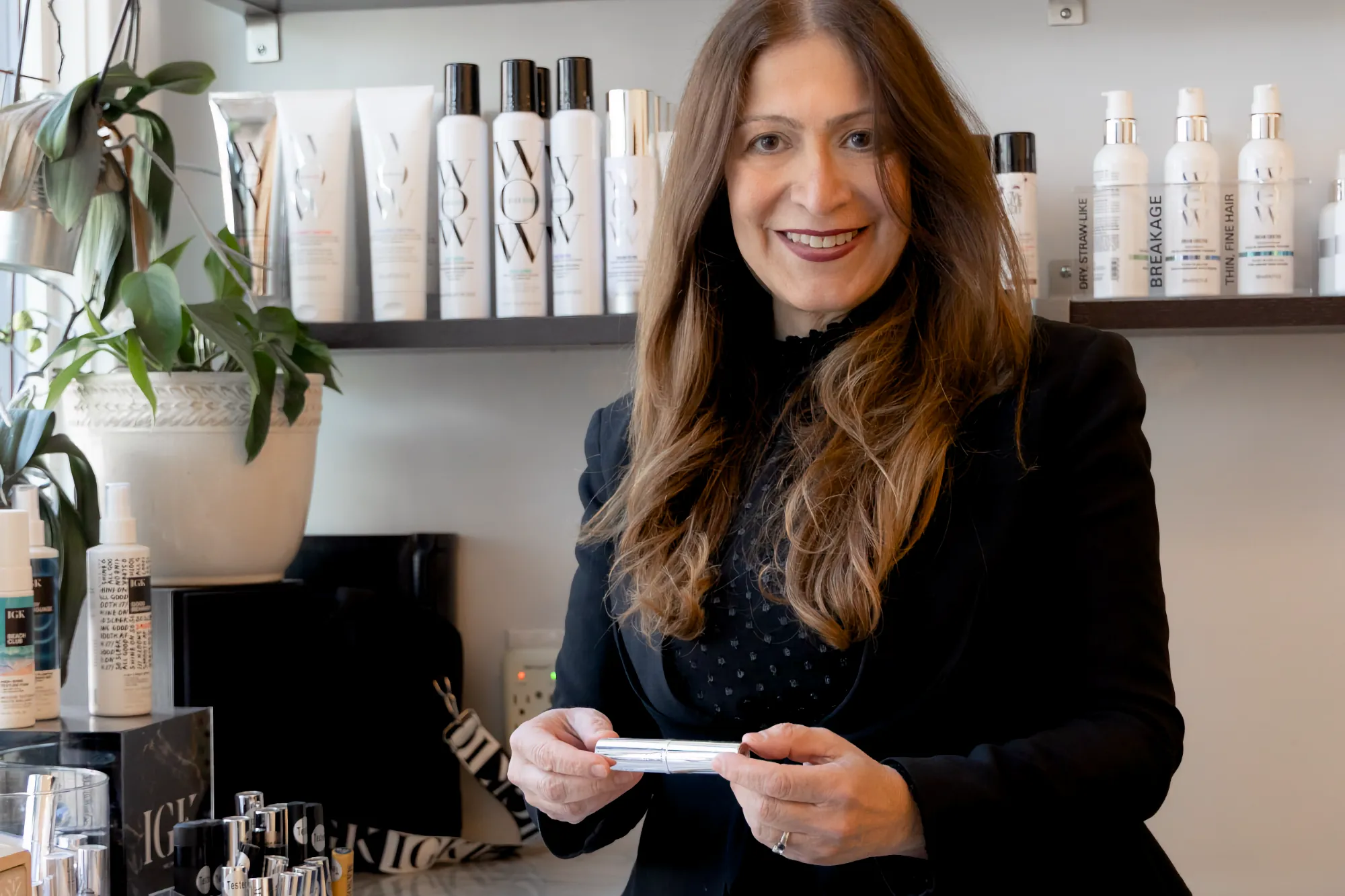 Salon owner Brigitte Rist smiles in her salon, holding a Manon product, with shelves of various beauty supplies behind her.