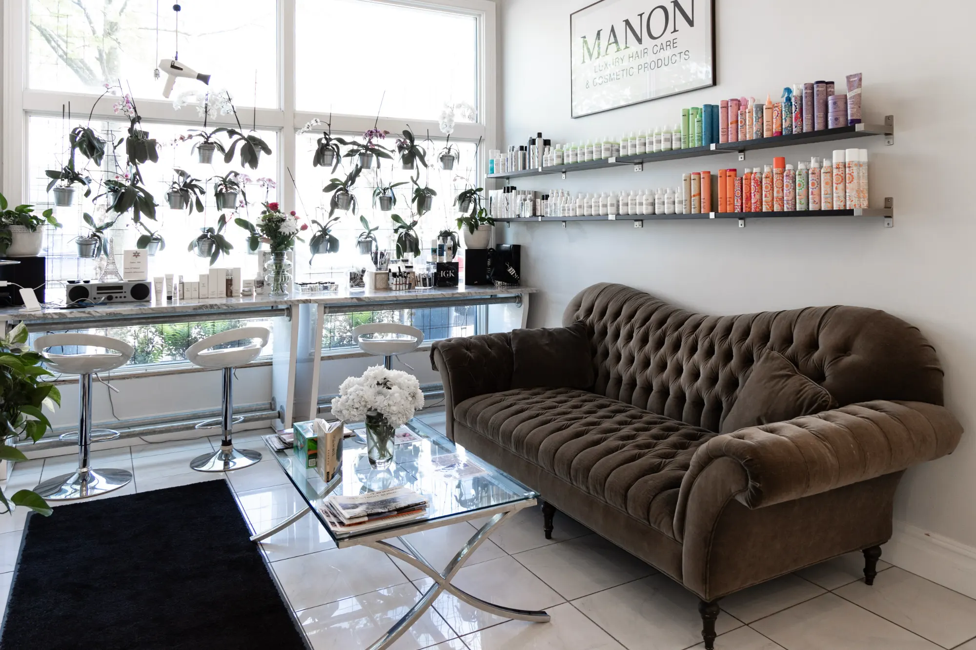 A luxurious salon waiting area featuring a brown velvet tufted sofa and a glass table with flowers. Shelves of beauty products line the white wall next to large windows adorned with orchids.