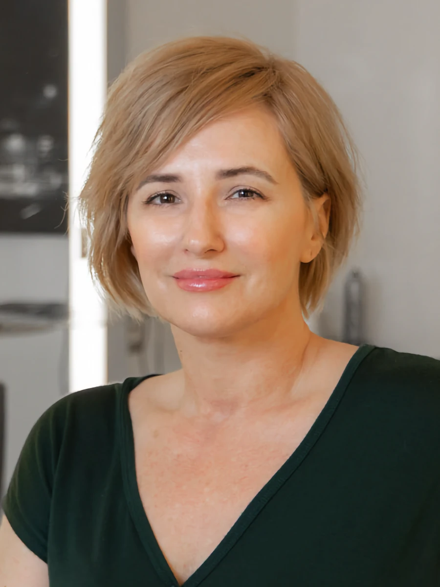 Misty Balido, an esthetician, with short blonde hair and a dark green top, offers a warm, gentle smile while looking directly at the camera.