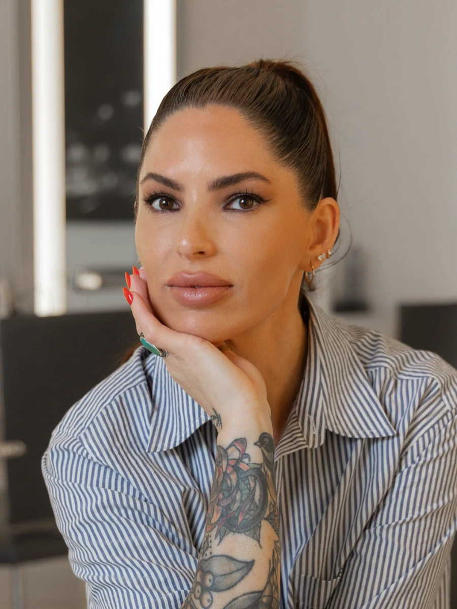 Lindsay Trudell, a permanent makeup artist, poses with a professional expression. She wears a striped shirt, rests her chin on her hand, and shows a full sleeve tattoo and red manicure.