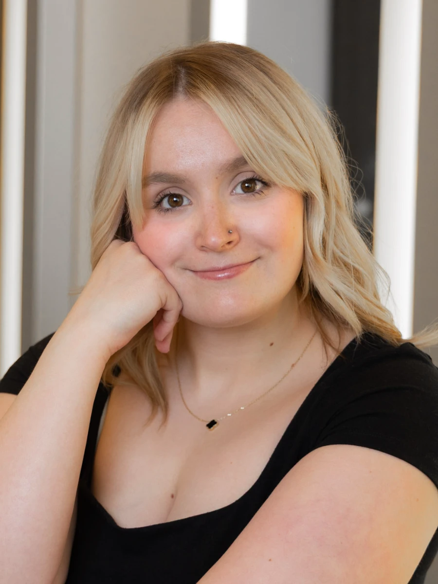 A blonde woman with a warm, friendly smile looks directly at the camera, resting her chin in her hand. She is wearing a black top and a gold necklace.