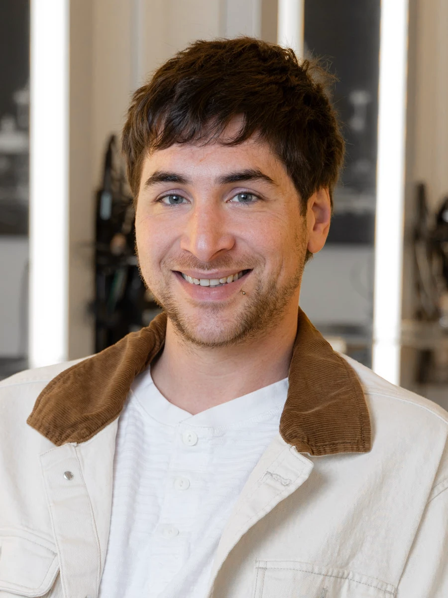 Stylist Matthew smiles warmly, wearing a white shirt and a light beige jacket with a brown corduroy collar. He has dark hair and a light goatee.