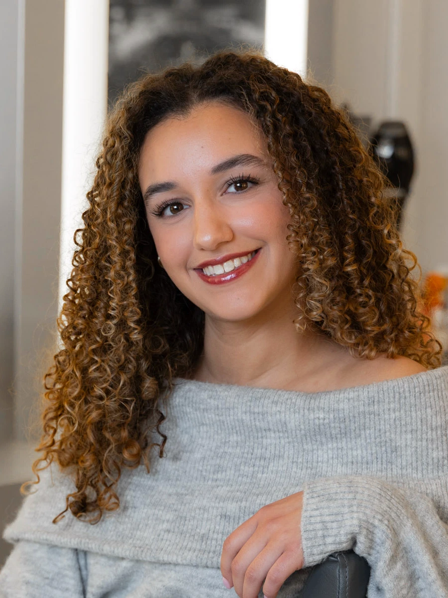 Olivia Hutchins, a stylist, with long curly brown hair and a gray sweater, smiles warmly at the camera.