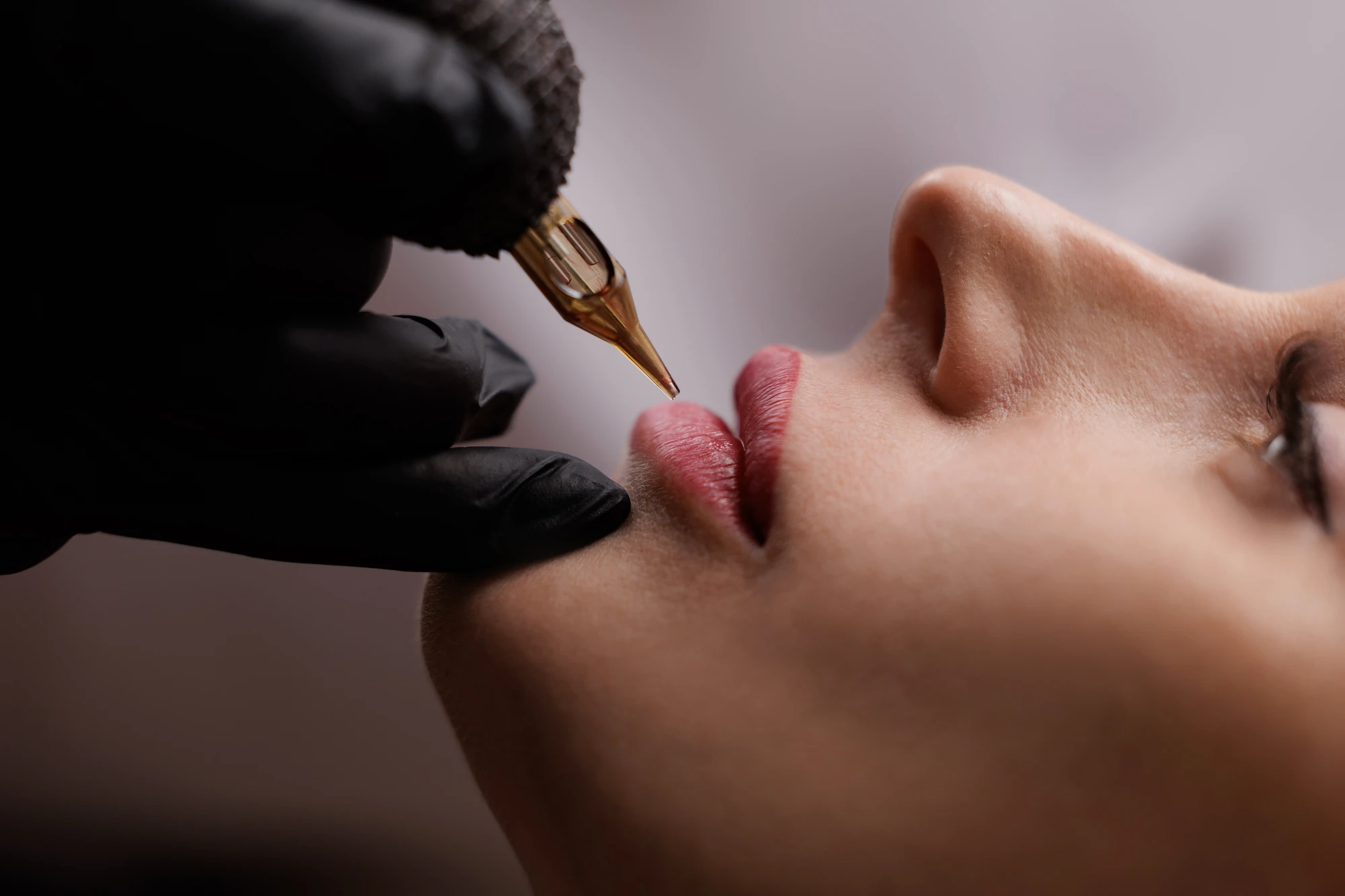 A gloved hand holds a cosmetic tattooing machine over a client's mouth during a lip pigmentation treatment.