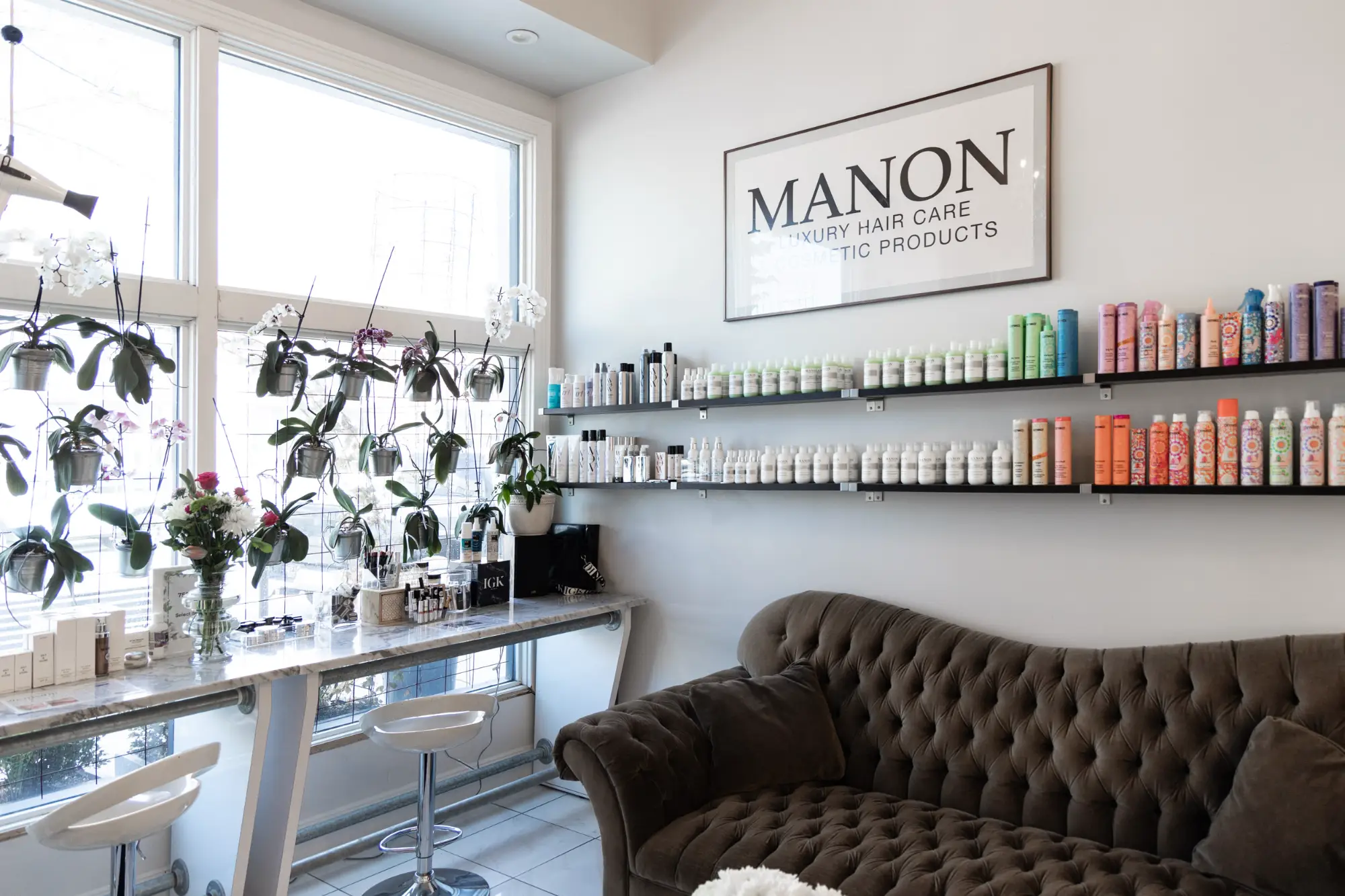 A bright salon waiting area features a dark tufted sofa, white product display shelves, and a marble counter near a window filled with hanging orchid plants.