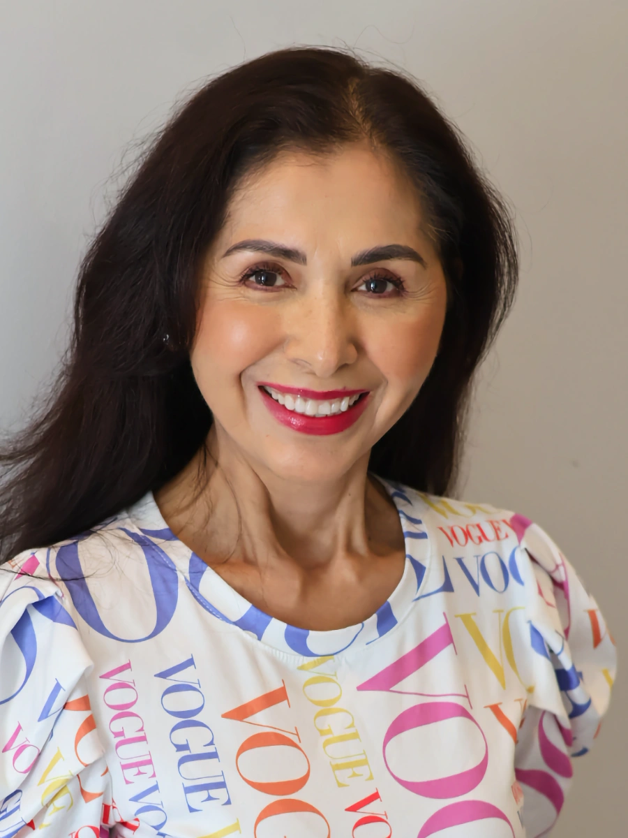 Myrna Perez, the Front Desk Coordinator, smiles broadly, wearing a white top with colorful lettering and vibrant red lipstick.