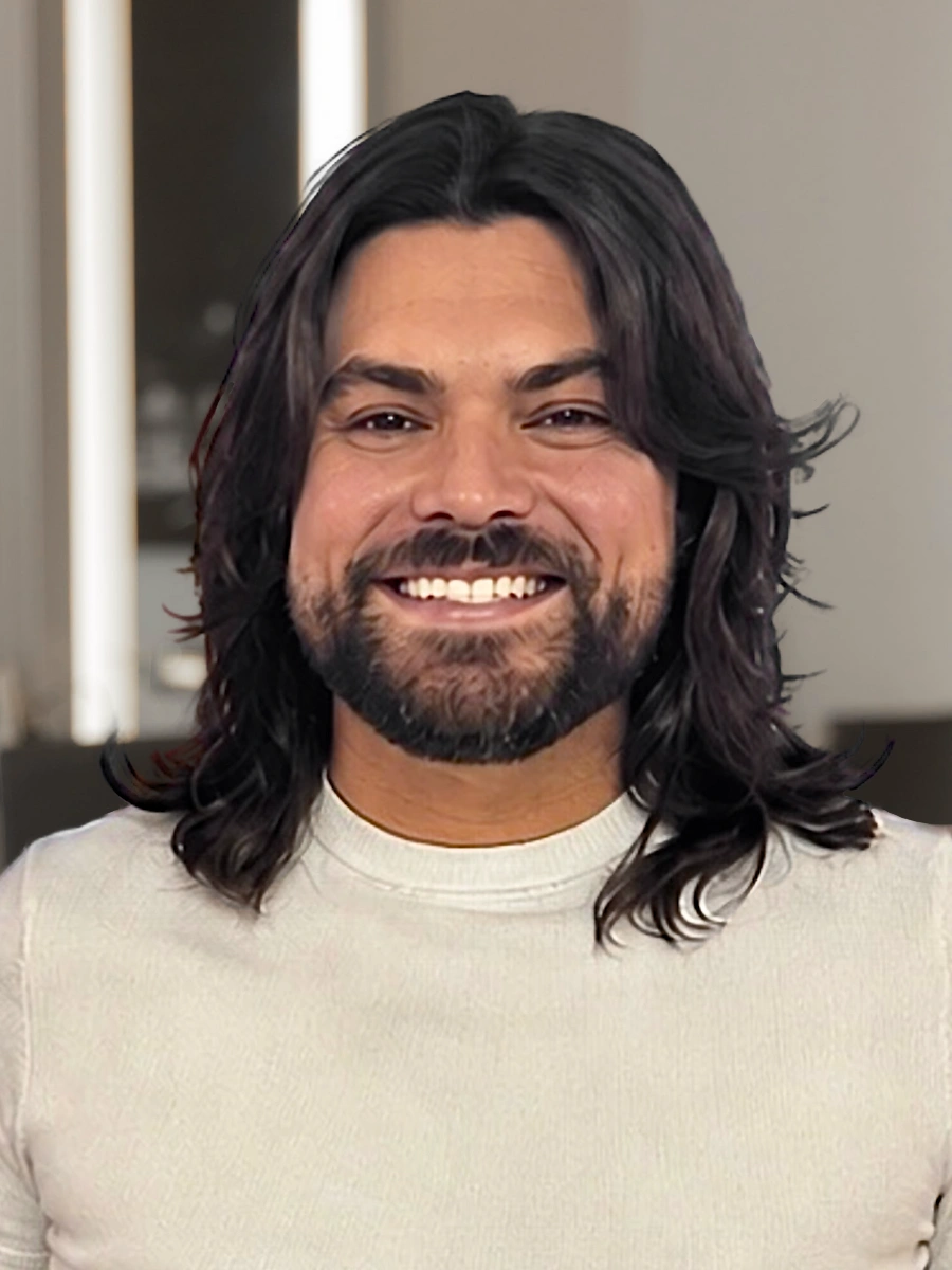 Michael Phillips, a hair stylist, smiles broadly, showing his teeth. He has long dark wavy hair and a beard, and wears a light-colored long-sleeved shirt.